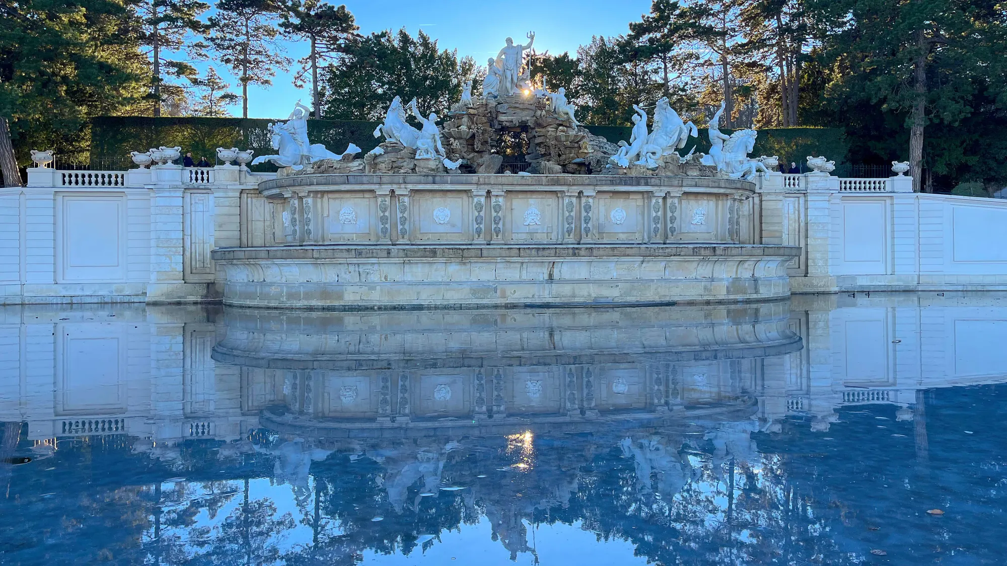 Marble fountain with the sun and trees set behind it and its reflection in the ice-covered pond in front of it