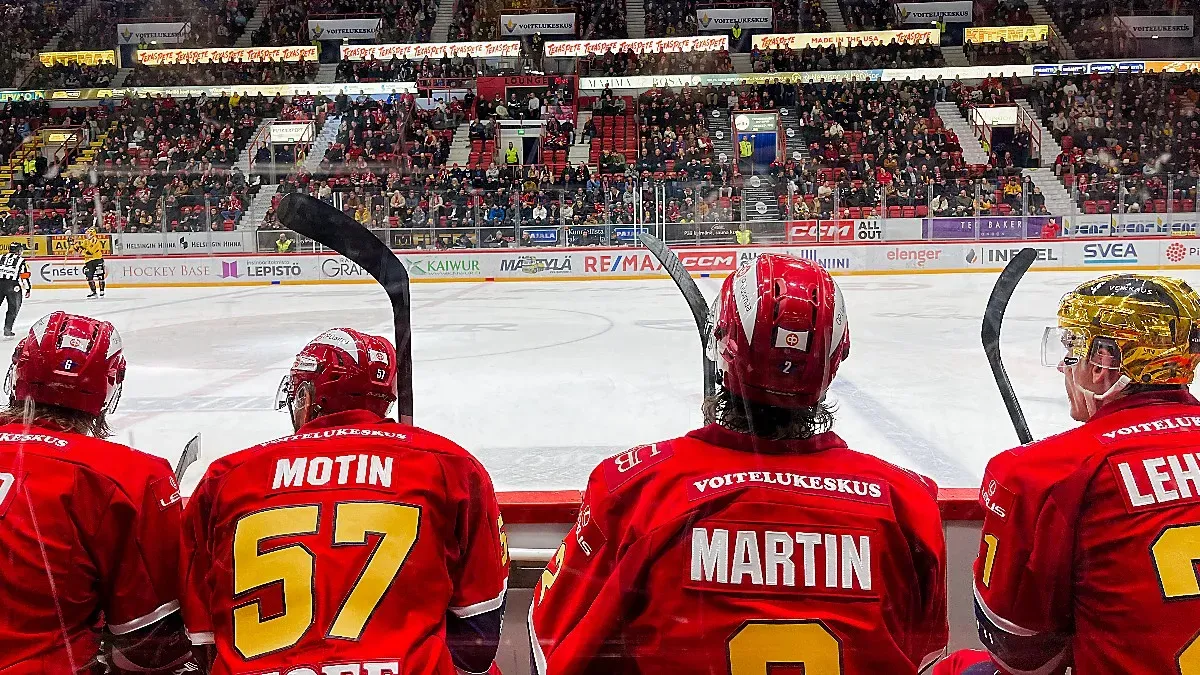 The backs of four hockey players sitting alongside an ice rink