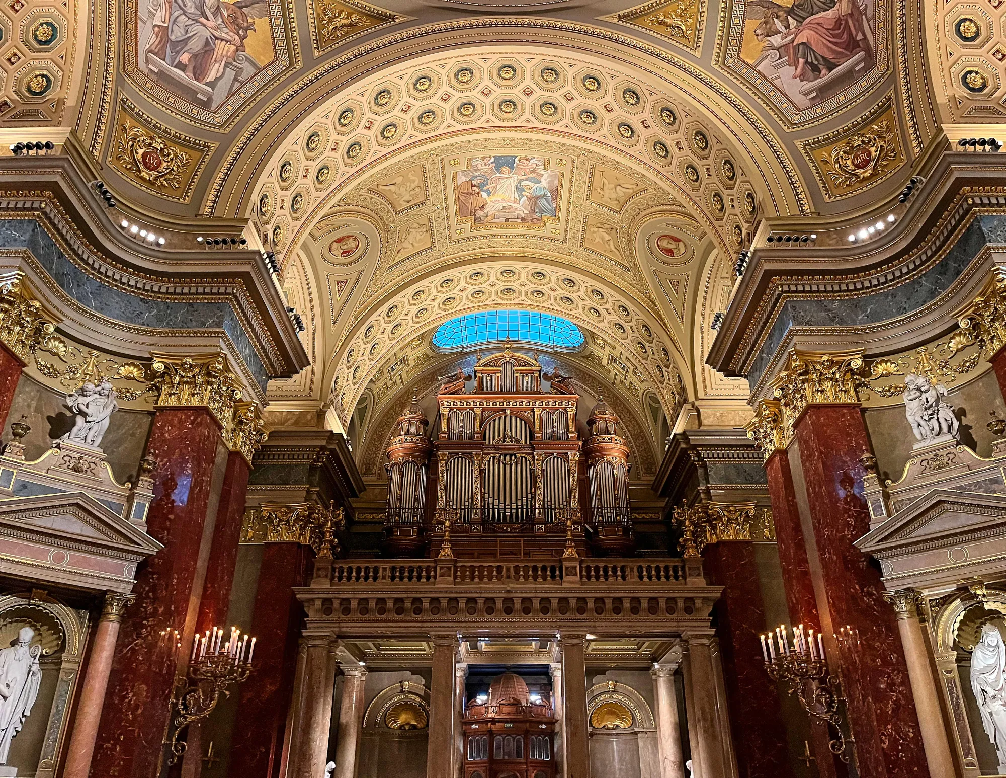 Red marble walls and ornate wood and gold organ under a golden ceilding with paintings and floral molding.