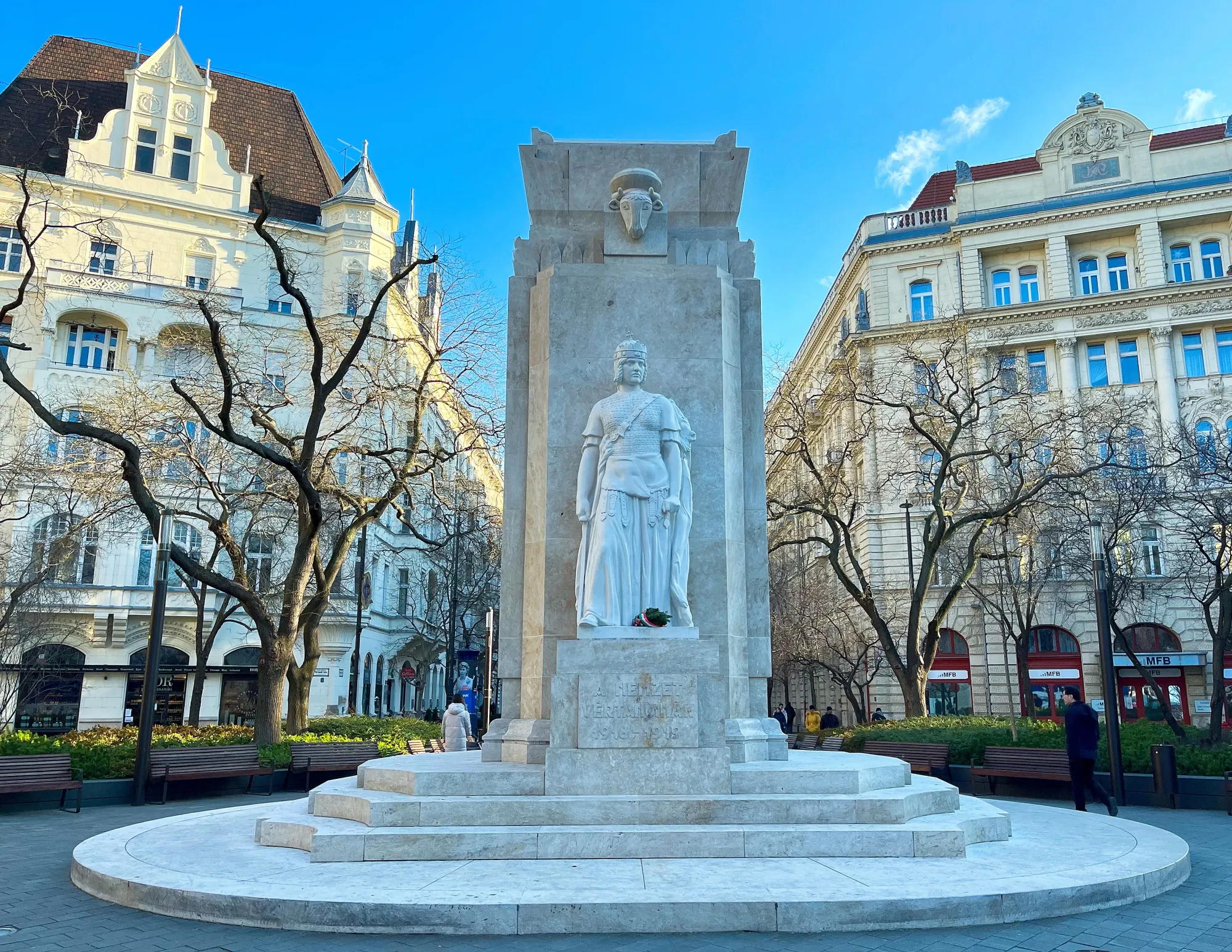 Stone rectangular column with a warrior on a pedastal resting in front of two cream-colored buildings