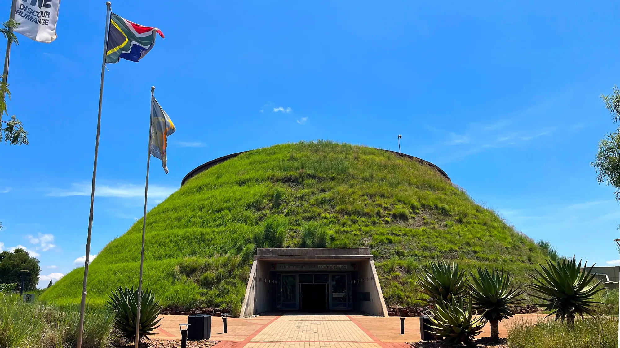 Grass covered conical building with a gaping entrance and adjoining flags