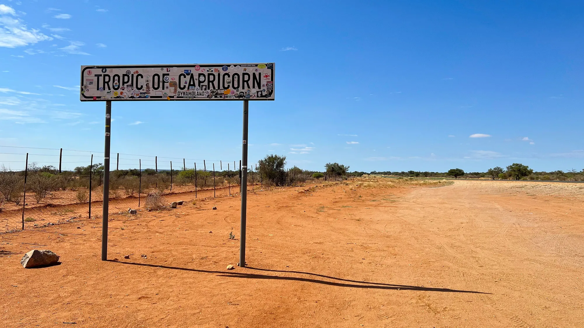 Flat sandy road with a white sign reading "Tropic of Capricorn" covered in stickers