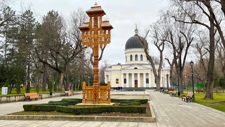 Wooden art piece imbedded in low-lying hedges with a cream-colored church set behind it.