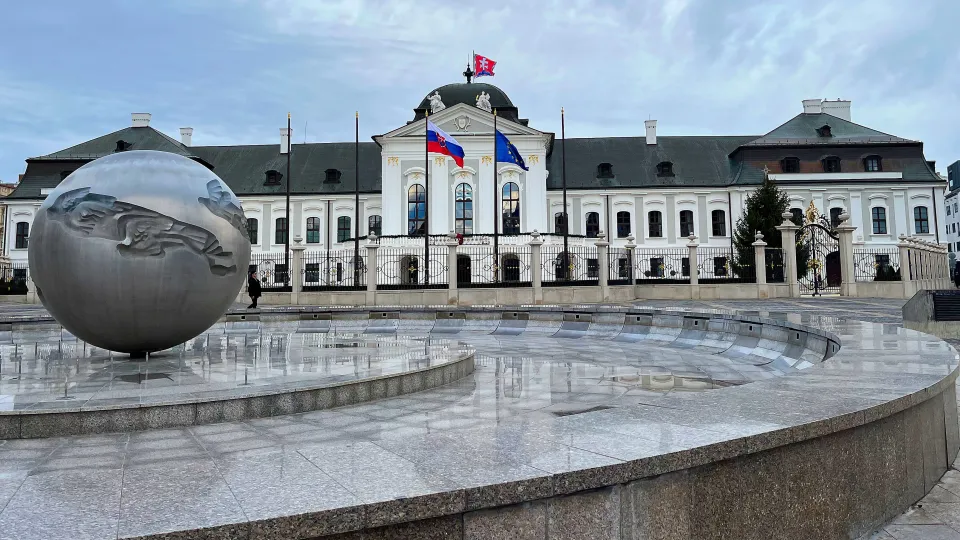 White elongated building with the Slovakian and European Union flags flying behind an orb shaped fountain